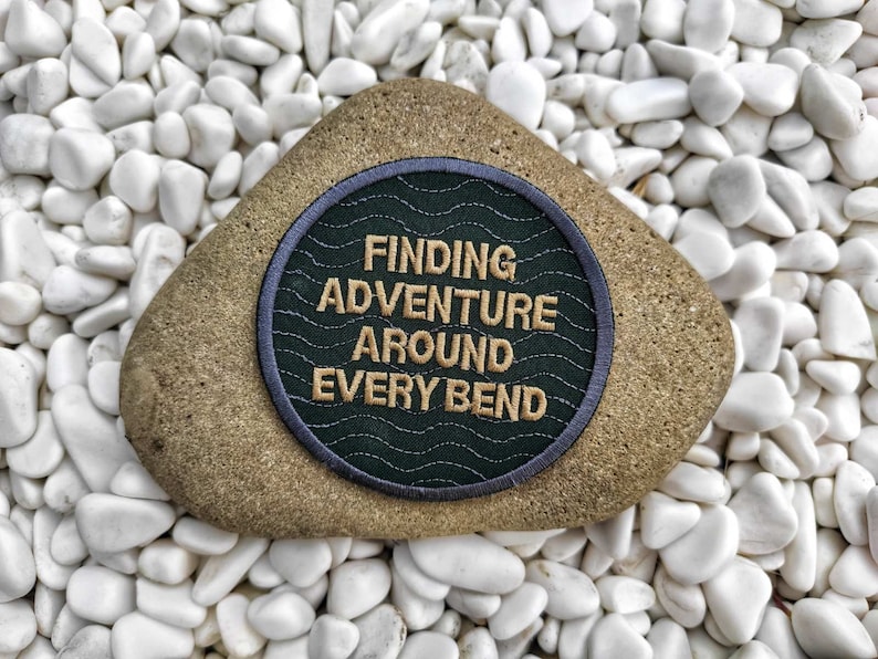 Peut inclure: Un patch brod&eacute; vert et gris avec le texte "FINDING ADVENTURE AROUND EVERY BEND" en fil dor&eacute;. Le patch est sur un rocher blanc.