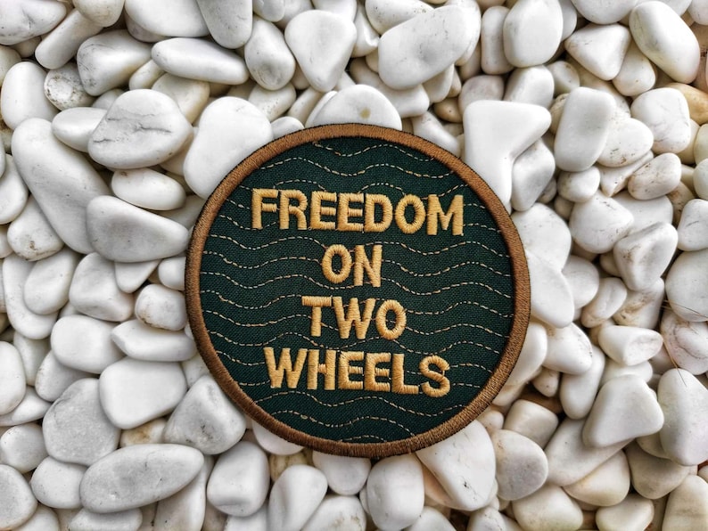 Peut inclure: Un &eacute;cusson brod&eacute; vert et or avec le texte "FREEDOM ON TWO WHEELS" sur un fond blanc de petites pierres.
