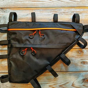 May include: Black bicycle frame bag with orange zipper accents and red zipper pulls. The bag has multiple straps for secure attachment and is designed to fit within the frame of a bicycle. The bag is made of a durable, water-resistant material.