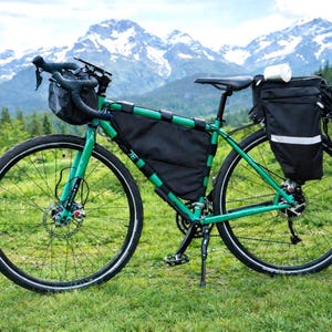 May include: A green bicycle with black storage bags and a water bottle, set against a backdrop of mountains and trees. The bike has black tires and a black seat. The frame is green with black accents.