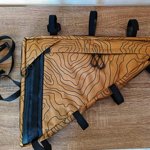 Pode incluir: Uma bolsa de quadro de bicicleta marrom e preta com um padrão topográfico. A bolsa tem várias correias e fivelas para prender a uma bicicleta.