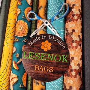Peut inclure: Gros plan sur des rouleaux de tissu avec diff&eacute;rents motifs, dont des champignons, des sapins et des motifs g&eacute;om&eacute;triques. Une paire de ciseaux est plac&eacute;e sur les rouleaux, et une pancarte en bois indique "Made in Ukraine LESENOK BAGS".