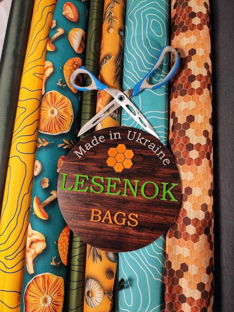 Peut inclure: Gros plan sur des rouleaux de tissu avec diff&eacute;rents motifs, dont des champignons, des sapins et des motifs g&eacute;om&eacute;triques. Une paire de ciseaux est plac&eacute;e sur les rouleaux, et une pancarte en bois indique "Made in Ukraine LESENOK BAGS".