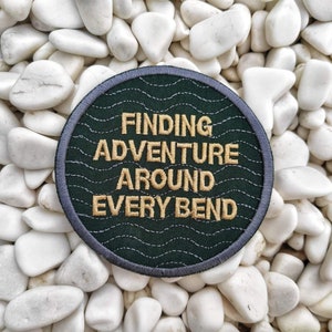 Peut inclure: Un &eacute;cusson brod&eacute; vert fonc&eacute; avec des points de couture blancs. L'&eacute;cusson porte le texte "Finding Adventure Around Every Bend" en fil dor&eacute;.