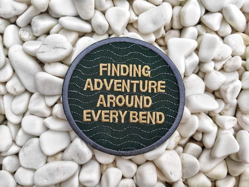 Peut inclure: Un &eacute;cusson brod&eacute; vert fonc&eacute; avec des points de couture blancs. L'&eacute;cusson porte le texte "Finding Adventure Around Every Bend" en fil dor&eacute;.