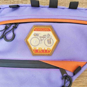 Peut inclure: Sac de v&eacute;lo violet et orange avec une fermeture &eacute;clair et un patch qui dit "Cycling Trip" avec une illustration de v&eacute;lo.