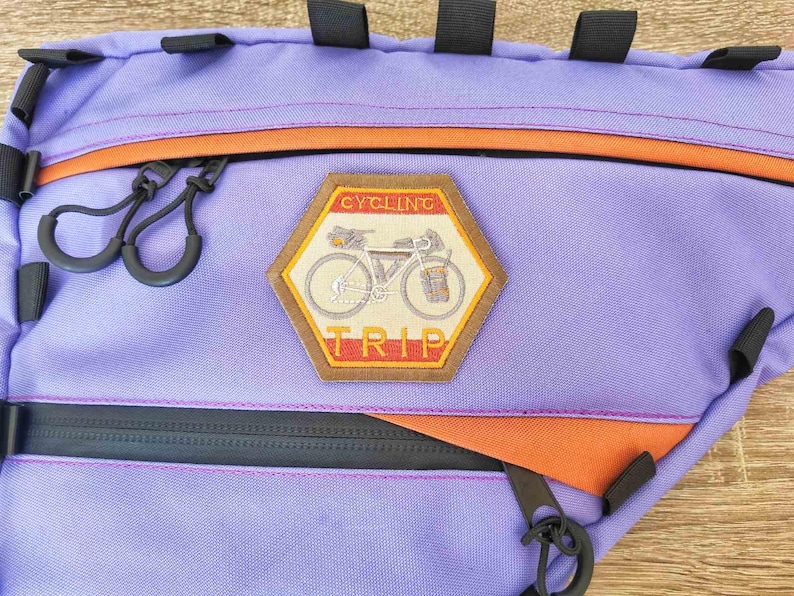Peut inclure: Sac de v&eacute;lo violet et orange avec une fermeture &eacute;clair et un patch qui dit "Cycling Trip" avec une illustration de v&eacute;lo.