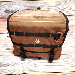 Könnte beinhalten: Braune Canvas-Tasche mit schwarzen Riemen und Schnallen. Die Tasche hat ein weißes Blumenmuster und einen braunen, tentakelartigen Verschluss. Die Tasche ist mit mehreren Fächern ausgestattet und eignet sich für Outdoor-Aktivitäten.