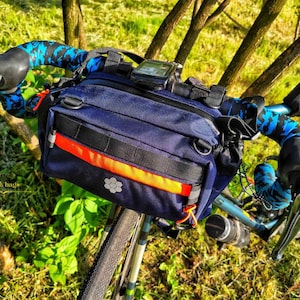 Może przedstawiać: Niebieska torba na kierownicę z jaskrawym pomarańczowym paskiem i czarnym zamkiem błyskawicznym. Torba jest przymocowana do kierownicy roweru za pomocą niebieskiej i czarnej taśmy na kierownicę.