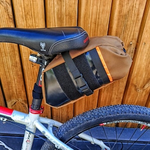 Puede incluir: Una bolsa de sillín de bicicleta de lona marrón con correas negras y cierre de velcro. La bolsa está unida al poste del sillín de la bicicleta.