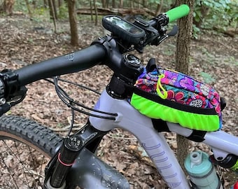 Op de afbeelding: Een grijze en zwarte mountainbike met een kleurrijke stuurtas en een zwart stuur. De fiets heeft een zwarte verende vork en een zwart-witte band. De fiets staat in een bosrijk gebied.