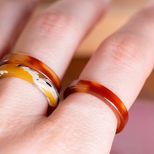 Op de afbeelding: Twee harsringen op een vinger. De ring op de wijsvinger is een enkele band met een bruin en oranje marmerpatroon. De ring op de middelvinger is een dubbele band met een geel en bruin marmerpatroon.