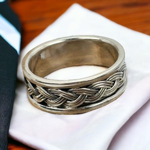 Könnte beinhalten: Ein silberner Ring mit einem geflochtenen Design. Der Ring liegt auf einer weißen Oberfläche.
