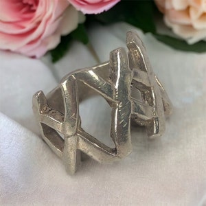 Op de afbeelding: Een zilveren ring met een geometrisch ontwerp. De ring is gemaakt van dik, getextureerd metaal en heeft een brede band.