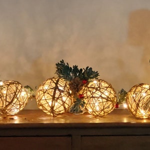 May include: A decorative display featuring spherical string lights. The spheres are crafted from woven brown material, housing small, warm white lights. Greenery and pinecones are arranged between the spheres, creating a festive display, perfect for the holiday season.