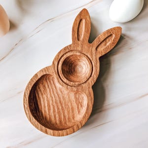 May include: A wooden bunny-shaped egg holder with a round indentation for holding an egg. The holder is made of light brown wood and has a natural wood finish.