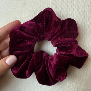 Puede incluir: Un coletero de terciopelo en color burdeos intenso con una textura suave y acanalada y un diseño circular fruncido. La tela tiene un tono rojo oscuro intenso que capta la luz, creando sutiles variaciones de color en los pliegues del accesorio.