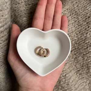 Puede incluir: Un par de pendientes de aro dorados con un acabado de esmalte marr&oacute;n claro, descansando en un plato blanco en forma de coraz&oacute;n.