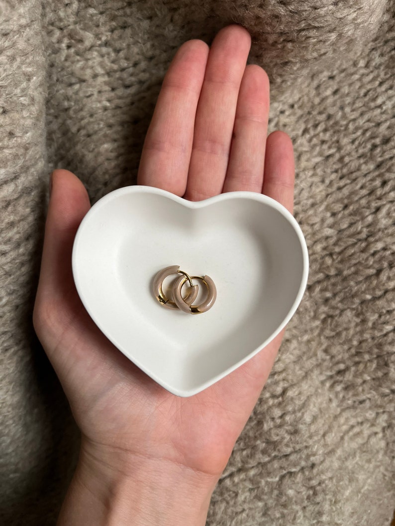 Puede incluir: Un par de pendientes de aro dorados con un acabado de esmalte marr&oacute;n claro, descansando en un plato blanco en forma de coraz&oacute;n.
