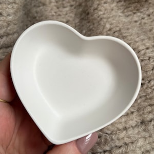 Puede incluir: Un plato blanco en forma de coraz&oacute;n hecho de silicona. El plato est&aacute; vac&iacute;o y est&aacute; siendo sostenido por una mano.