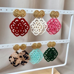 Puede incluir: Seis pares de pendientes colgantes con postes de metal martillado en tono dorado y colgantes geométricos calados. Los colgantes están hechos de resina acrílica en colores rojo, blanco, rosa, carey, azul claro y verde oscuro.