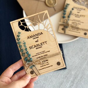 May include: A wooden wedding invitation with a laser cut design. The invitation is for Amanda Cruise and Scarlett Brown, and the wedding is on June 23, 2024 at the Garden in California. The invitation has a rustic feel with twine and dried flowers.