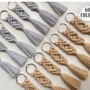 May include: A collection of handmade macrame keychains with tassels in grey and beige. The keychains feature a diamond pattern and are attached to silver key rings. The image includes the text "60 COLORS".