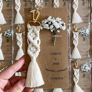 Peut inclure: Un porte-clés macramé fait à la main, avec un fermoir doré et une gland. Le porte-clés est associé à une petite carte avec les noms "Carlos & Ana" et la date "25/06/2021". Un petit bouquet de fleurs blanches est attaché à la carte.