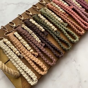 May include: A collection of personalised keychains with names spelled out on wooden beads. The keychains are made of braided rope in various shades of brown, beige, and green.