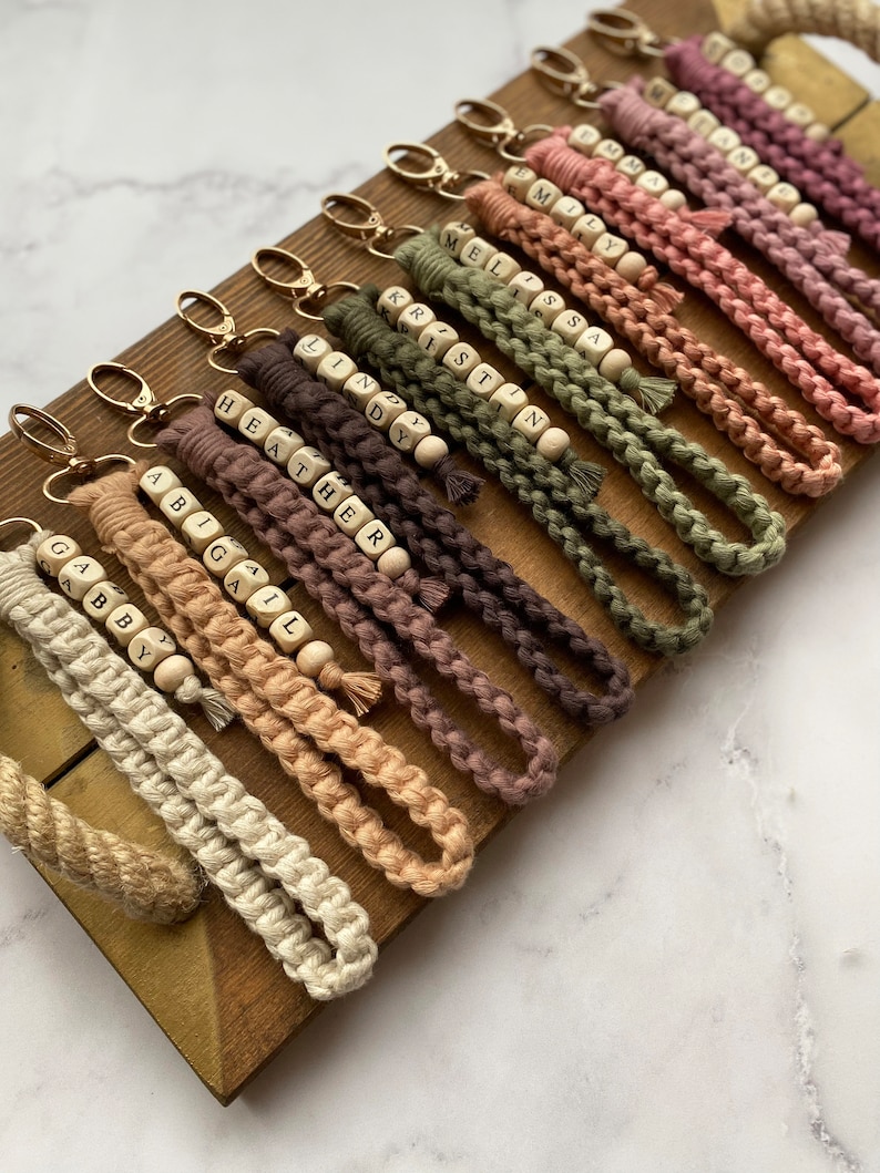 May include: A collection of personalised keychains with names spelled out on wooden beads. The keychains are made of braided rope in various shades of brown, beige, and green.
