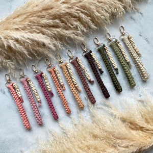 May include: A collection of 12 colourful keychains with personalised wooden beads spelling out different names. The keychains are made of macrame cord and have a gold metal ring.