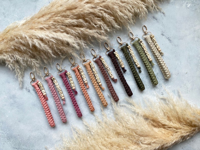 May include: A collection of 12 colourful keychains with personalised wooden beads spelling out different names. The keychains are made of macrame cord and have a gold metal ring.