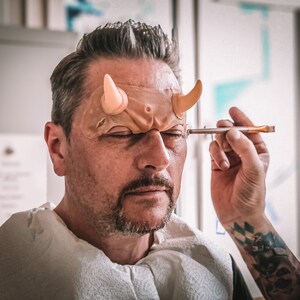 May include: A person is applying makeup to a man's forehead. The man is wearing a prosthetic with horns attached to it. The prosthetic is flesh-colored and the horns are a light peach color.