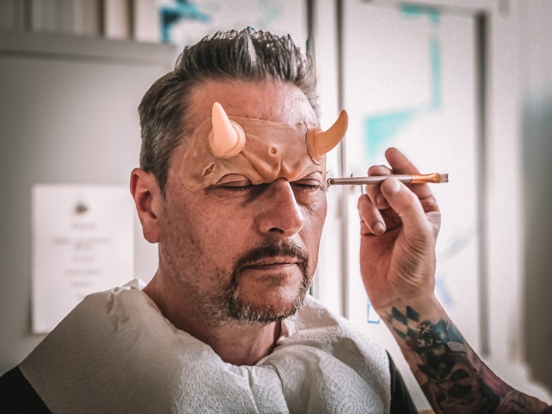 May include: A person is applying makeup to a man's forehead. The man is wearing a prosthetic with horns attached to it. The prosthetic is flesh-colored and the horns are a light peach color.