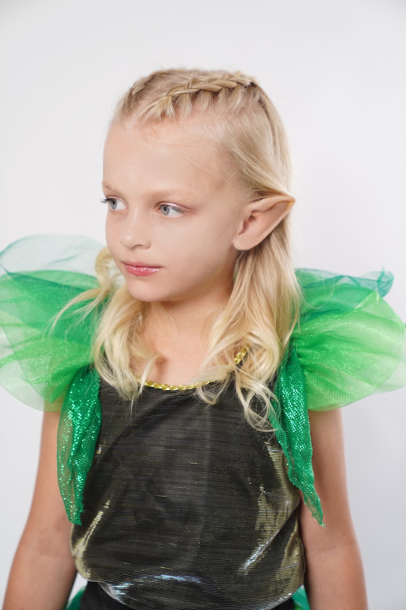 May include: A young girl wearing a green and gold costume with pointed ears. She has blonde hair braided across the top of her head.