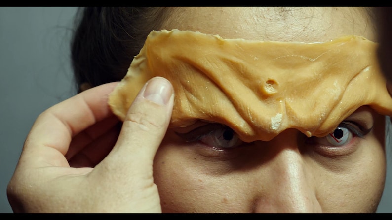 May include: A close-up of a person's face with a flesh-colored prosthetic covering one eye. The prosthetic is textured and has a small hole in it. The person is wearing white contact lenses.