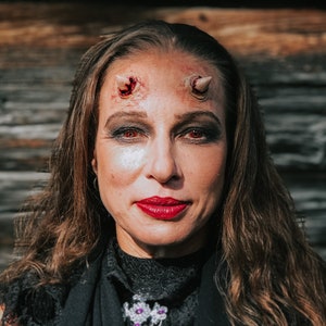 May include: A woman with long brown hair is wearing a black lace top and has red lipstick. She has fake horns on her forehead with fake blood dripping down her face.