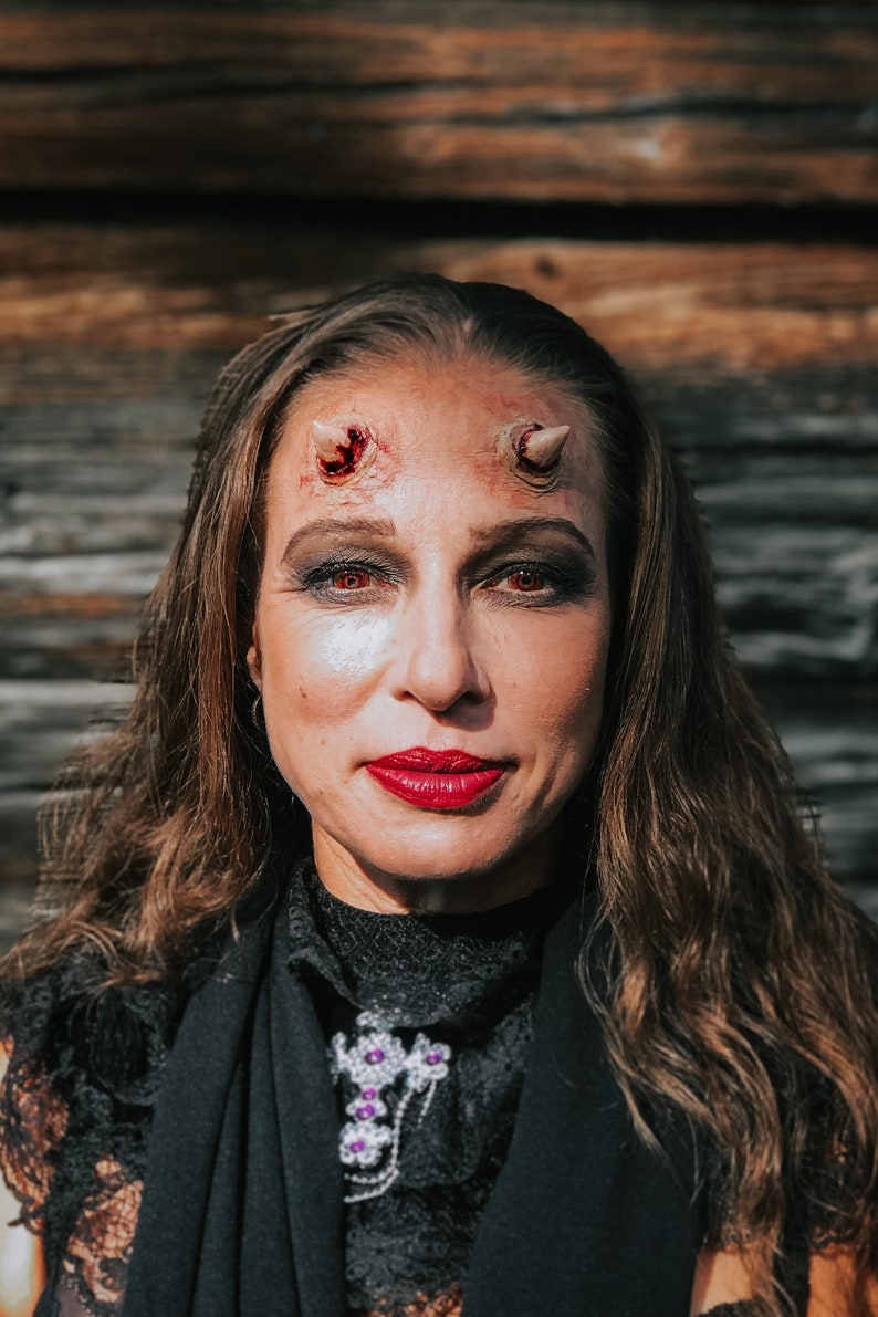 May include: A woman with long brown hair is wearing a black lace top and has red lipstick. She has fake horns on her forehead with fake blood dripping down her face.