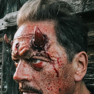 May include: A man with a bloody face and red devil horns on his forehead. The man is looking to the left of the frame.