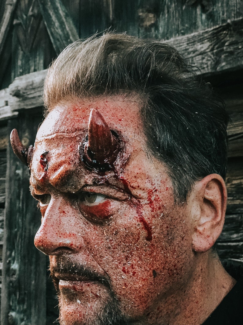 May include: A man with a bloody face and red devil horns on his forehead. The man is looking to the left of the frame.