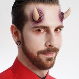 Puede incluir: Una persona con maquillaje temático de diablo, con cuernos protésicos y lentes de contacto rojos. La persona lleva una camisa roja con cuello y una corbata negra. Los cuernos son de color beige claro, y el maquillaje incluye heridas simuladas.