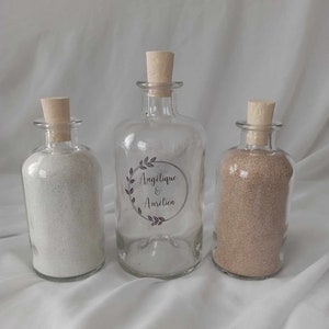 Peut inclure: Trois bouteilles en verre avec des bouchons en liège remplies de sable. La bouteille du milieu porte une étiquette avec le texte "Angelique & Aurelien" dans une police décorative.