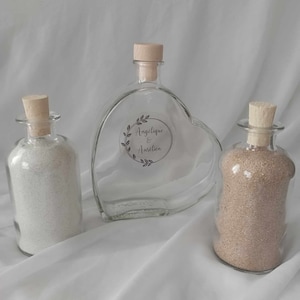 Peut inclure: Deux bouteilles en verre transparent avec des bouchons en liège remplies de sable, et une plus grande bouteille en verre en forme de cœur avec un bouchon en liège. La bouteille en forme de cœur porte une étiquette sur laquelle est inscrit "Angélique & Aurélien".