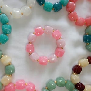 Peut inclure: Une collection de bracelets circulaires composés de perles en résine lisses et carrées de différentes couleurs, notamment rose, blanc, bleu sarcelle, vert et rouge foncé. Chaque perle est séparée par une petite perle intercalaire dorée.