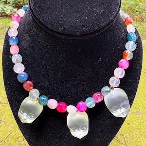 May include: A vibrant necklace with multi-coloured gemstone beads, including pink, blue, and clear stones. The necklace features three translucent doll-face pendants. It is displayed on a black velvet bust.