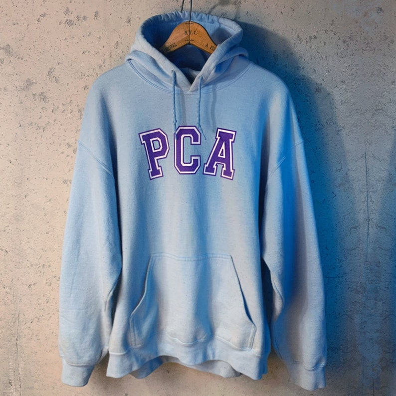 May include: A light blue hooded sweatshirt with a purple and white "PCA" logo on the front.