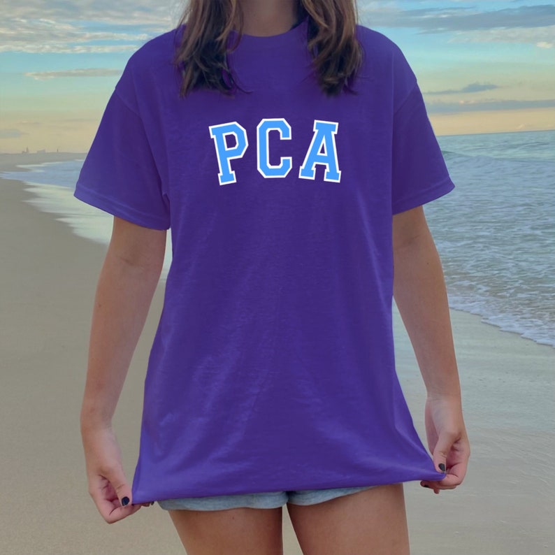 K&ouml;nnte beinhalten: Ein lila T-Shirt mit dem Text "PCA" in blauen Buchstaben.