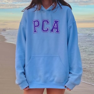 May include: A light blue hoodie with a purple "PCA" logo on the front. The hoodie has a drawstring hood and a kangaroo pocket.