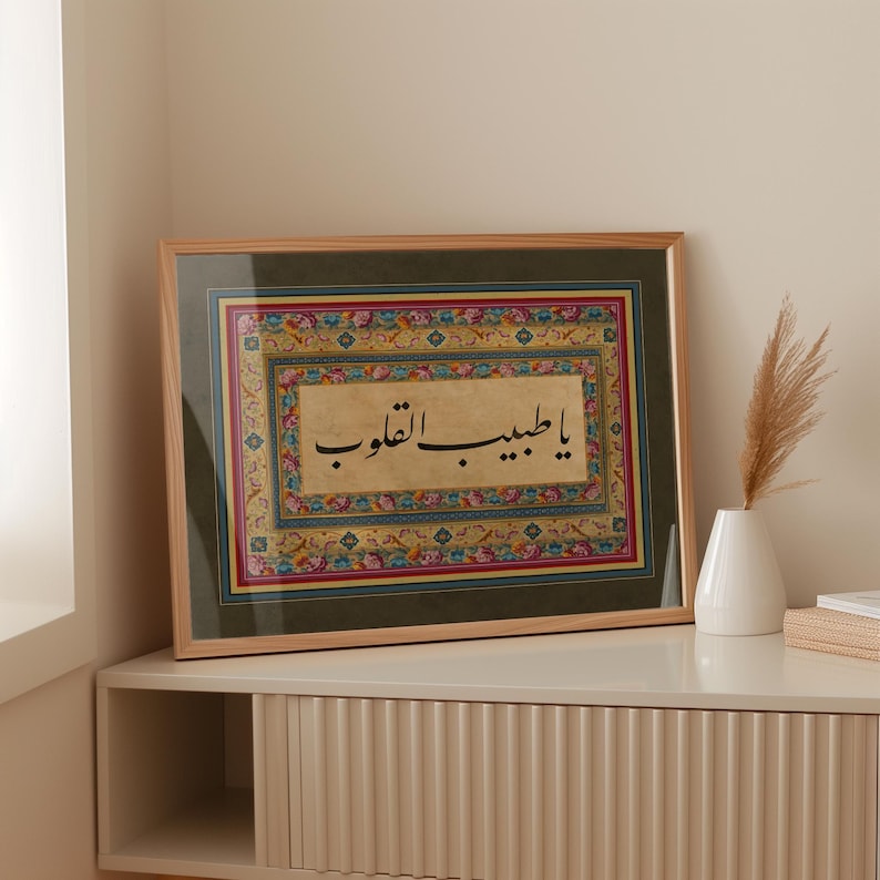May include: A framed artwork with a decorative border featuring intricate floral patterns in shades of red, blue, and gold. The artwork features a calligraphic inscription in black ink on a beige background. The inscription reads "يا طيب القلب" in Arabic.