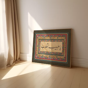 May include: A framed print with a gold frame and a green mat. The print features a decorative border with floral motifs in shades of pink, blue, and yellow. The center of the print features Arabic calligraphy in black ink on a cream background. The text reads "Ya Habibi El A'la".